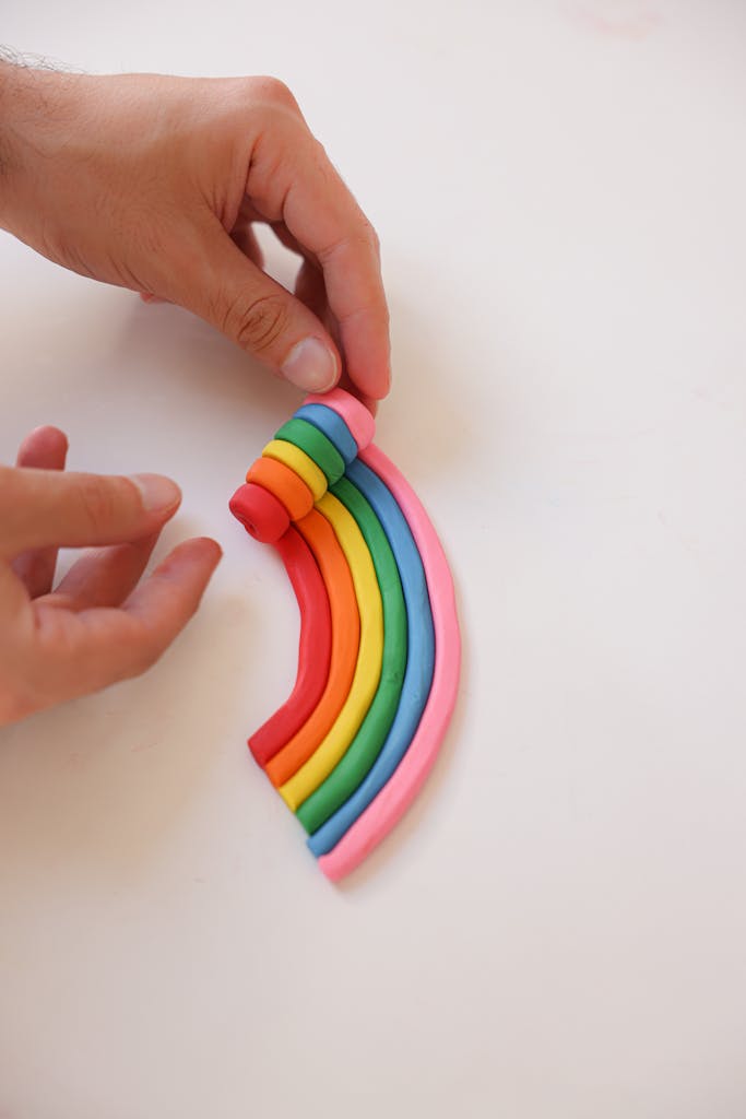 Hands shaping a vibrant clay rainbow on a white surface, showcasing creativity.